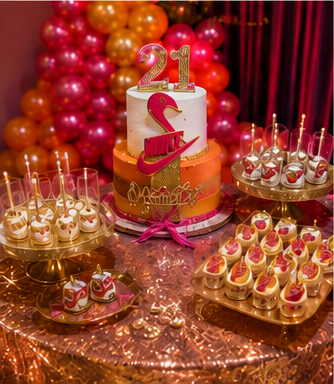 A birthday celebration table with a cake featuring the number 21, surrounded by pink, orange, and red balloons, and decorated with pink ribbons and gold accents. The table has various cupcakes and desserts with pink, white, and gold decorations.