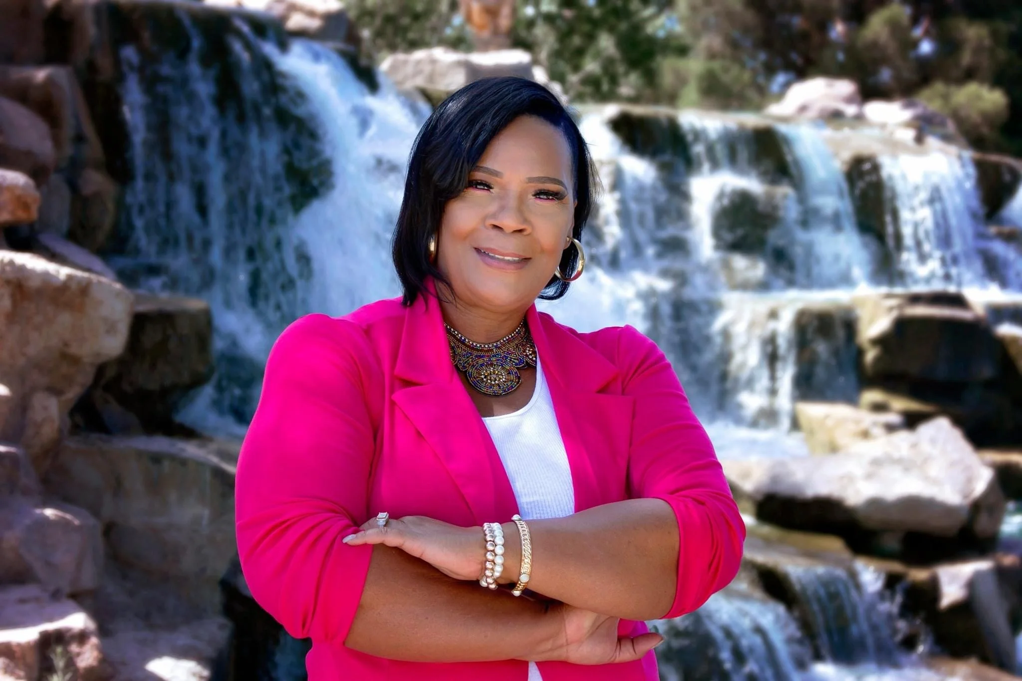 A woman with dark hair, wearing a pink blazer, jewelry, and crosses her arms, standing outdoors near a waterfall and rocks.
