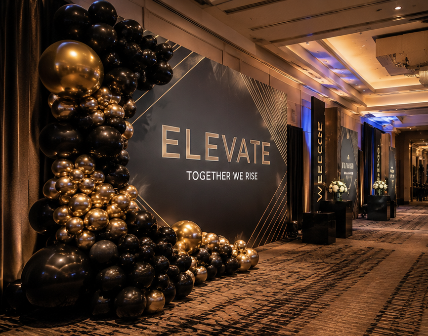 Elegant event space with a large black and gold balloon arrangement next to a backdrop that reads 'ELEVATE' and 'Together We Rise,' featuring black tables with white floral arrangements and a corridor with warm lighting and patterned carpet.