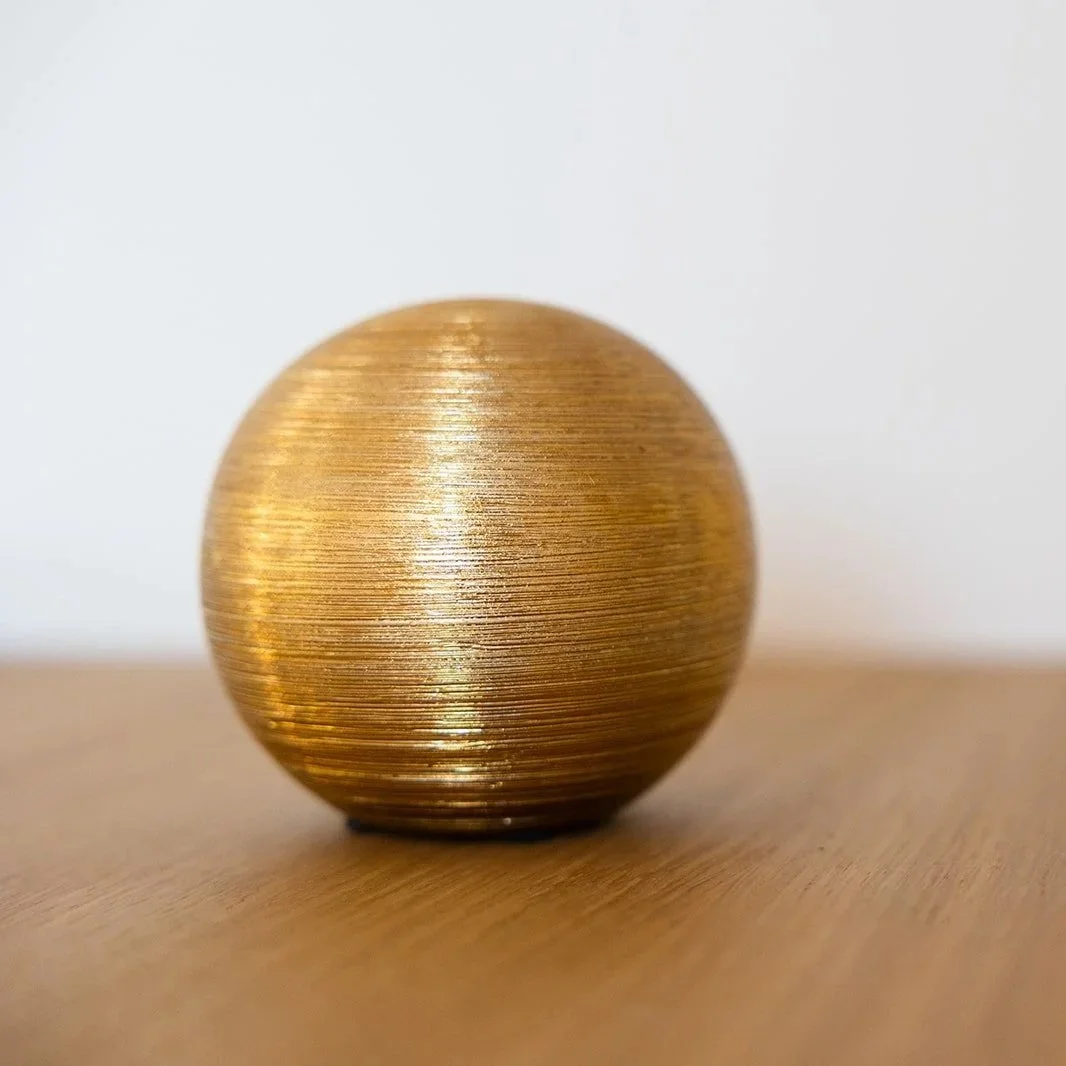 A gold-colored spherical object with a textured, brushed surface, placed on a wooden surface against a plain white background.