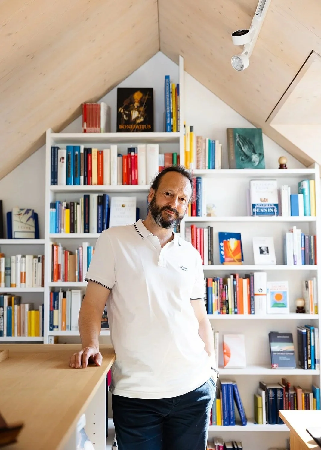 A man with dark hair and a beard standing in front of bookshelves filled with colorful books in a room with a sloped wooden ceiling.
