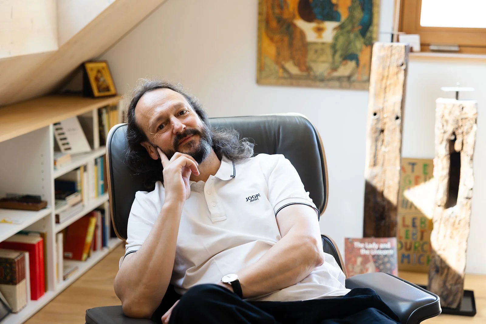 A man with long hair and a beard sitting in a black leather chair in a room with wooden shelves filled with books, artwork on the wall, and carved wooden sculptures in the background.