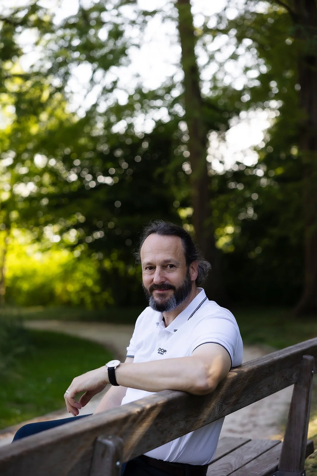 A man with a beard and mustache, wearing a white polo shirt, sitting on a wooden park bench outdoors with trees and greenery in the background, smiling at the camera.