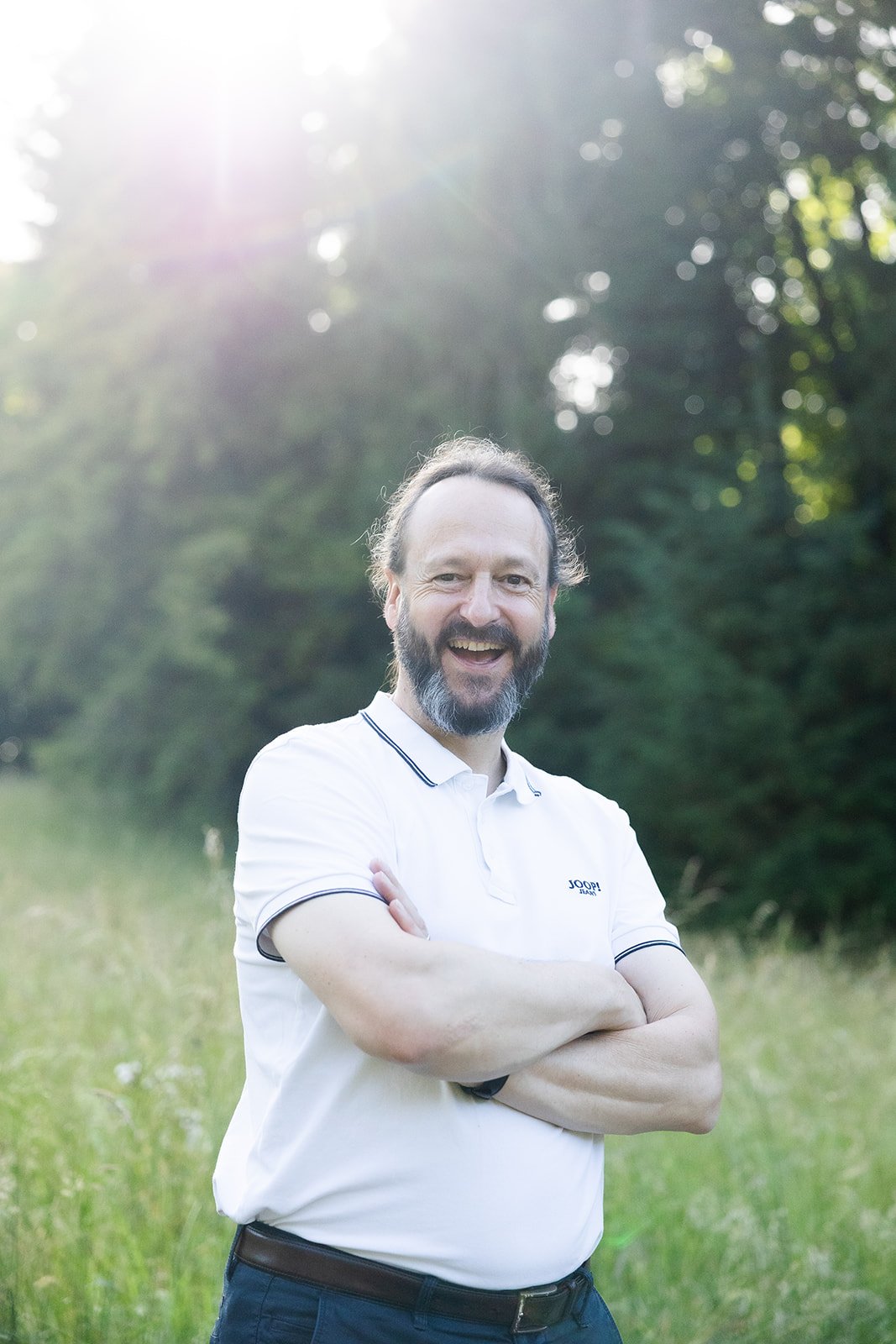 A man with a beard and long hair smiling with arms crossed outdoors in a grassy field, backlit by sunlight through trees.