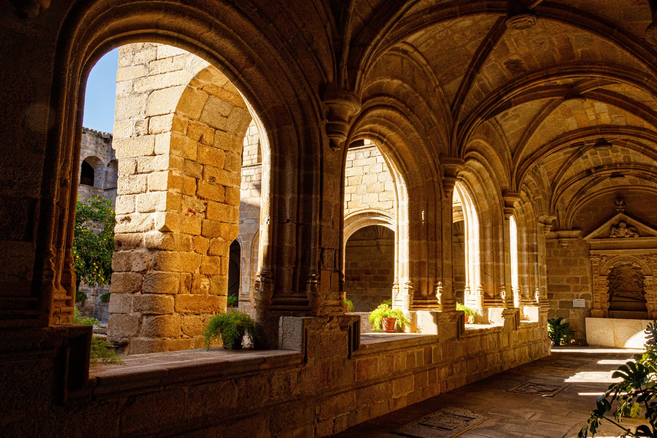 Sunlit stone arches and columns inside an ancient church or monastery courtyard with plants and a blue sky visible through openings.