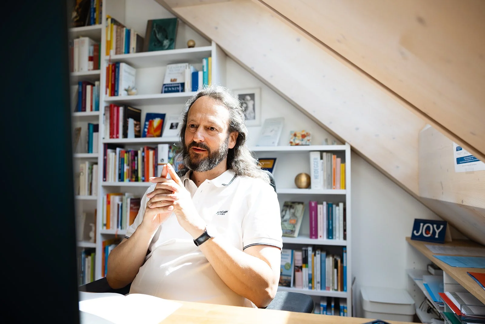A middle-aged man with long gray hair and beard sitting in a modern home office, wearing a white polo shirt, with shelves filled with books behind him and natural light coming from a window.