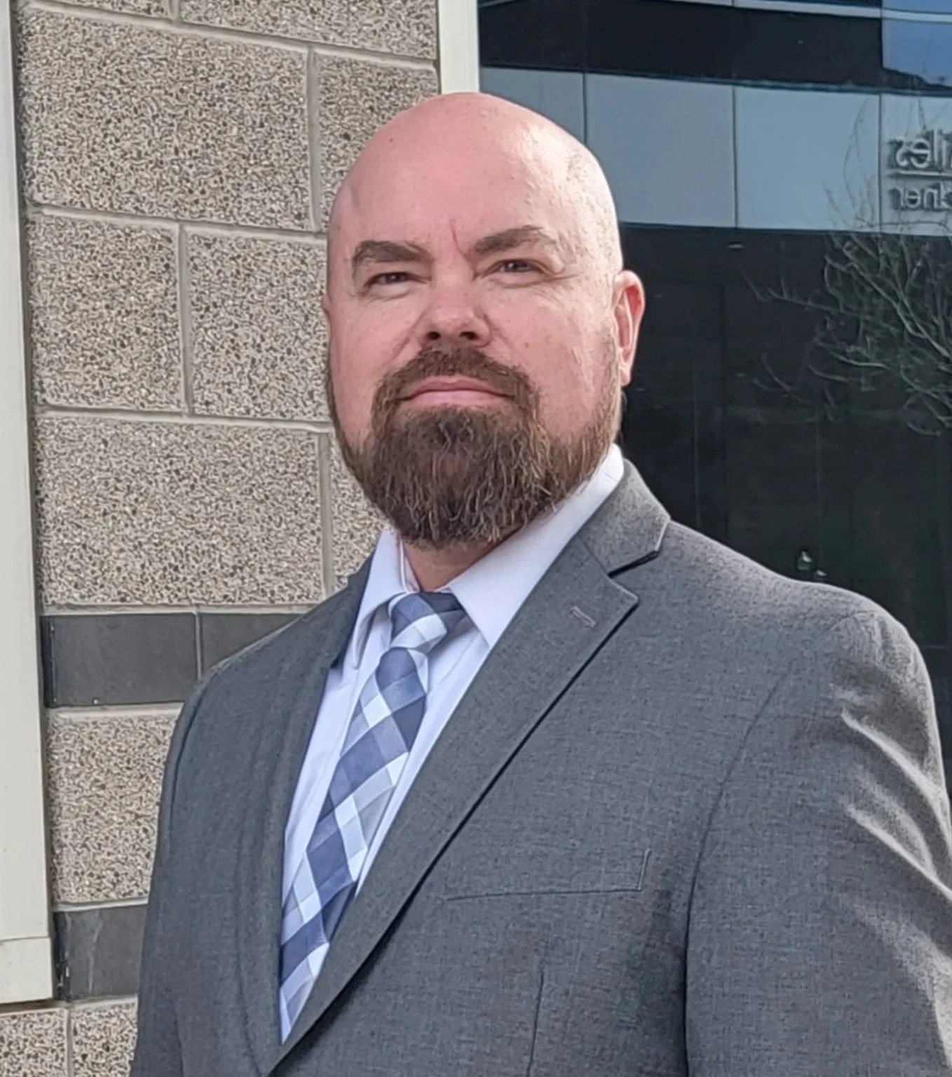 A man with a bald head and beard dressed in a gray suit with a white shirt and patterned tie standing outside near a building with textured brick and glass windows.