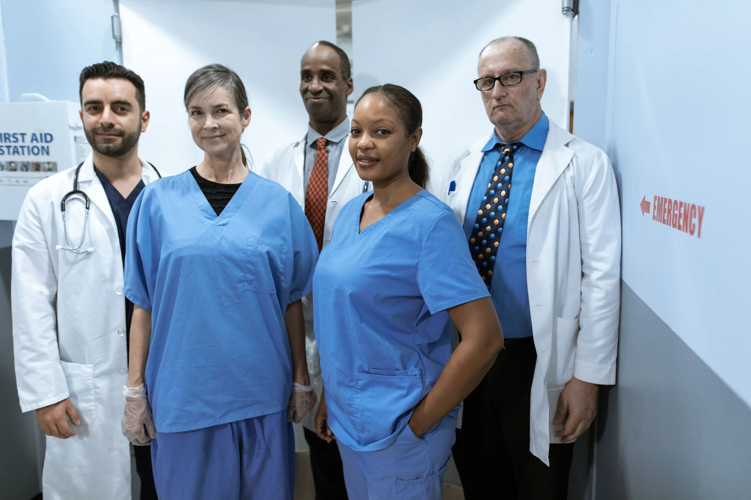 Group of five healthcare professionals, including nurses and doctors, standing together in a hospital hallway near an emergency sign and a first aid station.