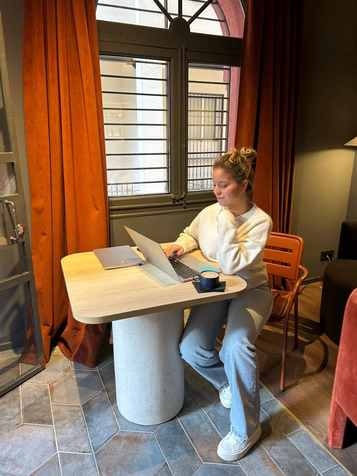 Beth sentada en una mesa de color claro con una computadora portátil, con una taza en la mano, en un ambiente interior con cortinas naranjas y ventanas con rejas.
