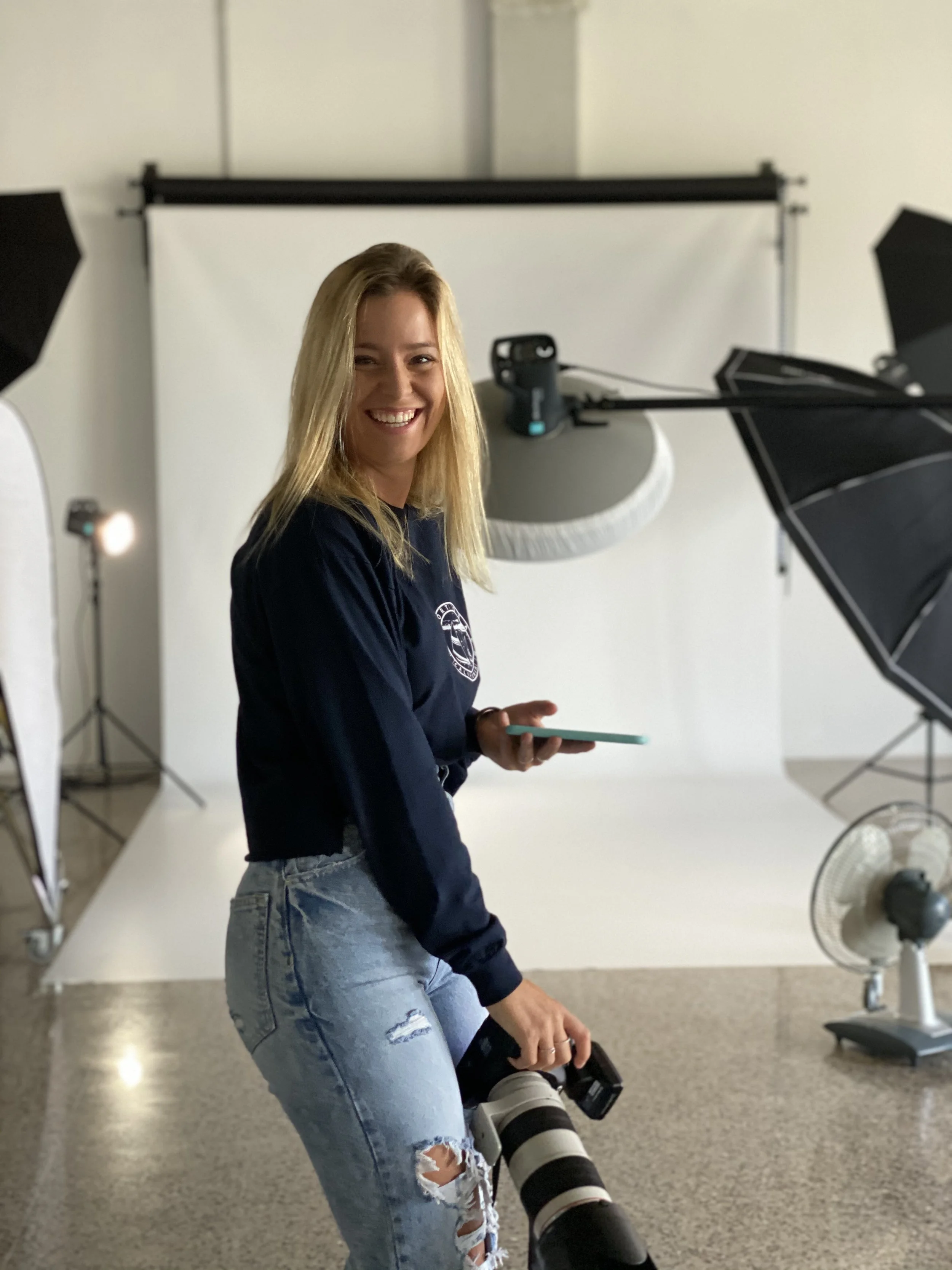 Una mujer sonriente con cabello rubio, usando una sudadera azul y jeans rotos, sosteniendo una cámara con teleobjetivo, en un estudio fotográfico con equipo de iluminación y fondo blanco.