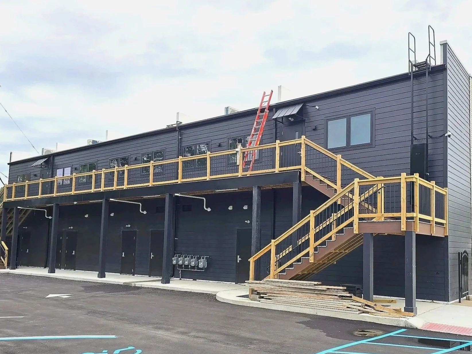 A two-story building with a black exterior, a wooden staircase, and a balcony. Ladder leaning against the building, parking lot with handicap spaces in the foreground.