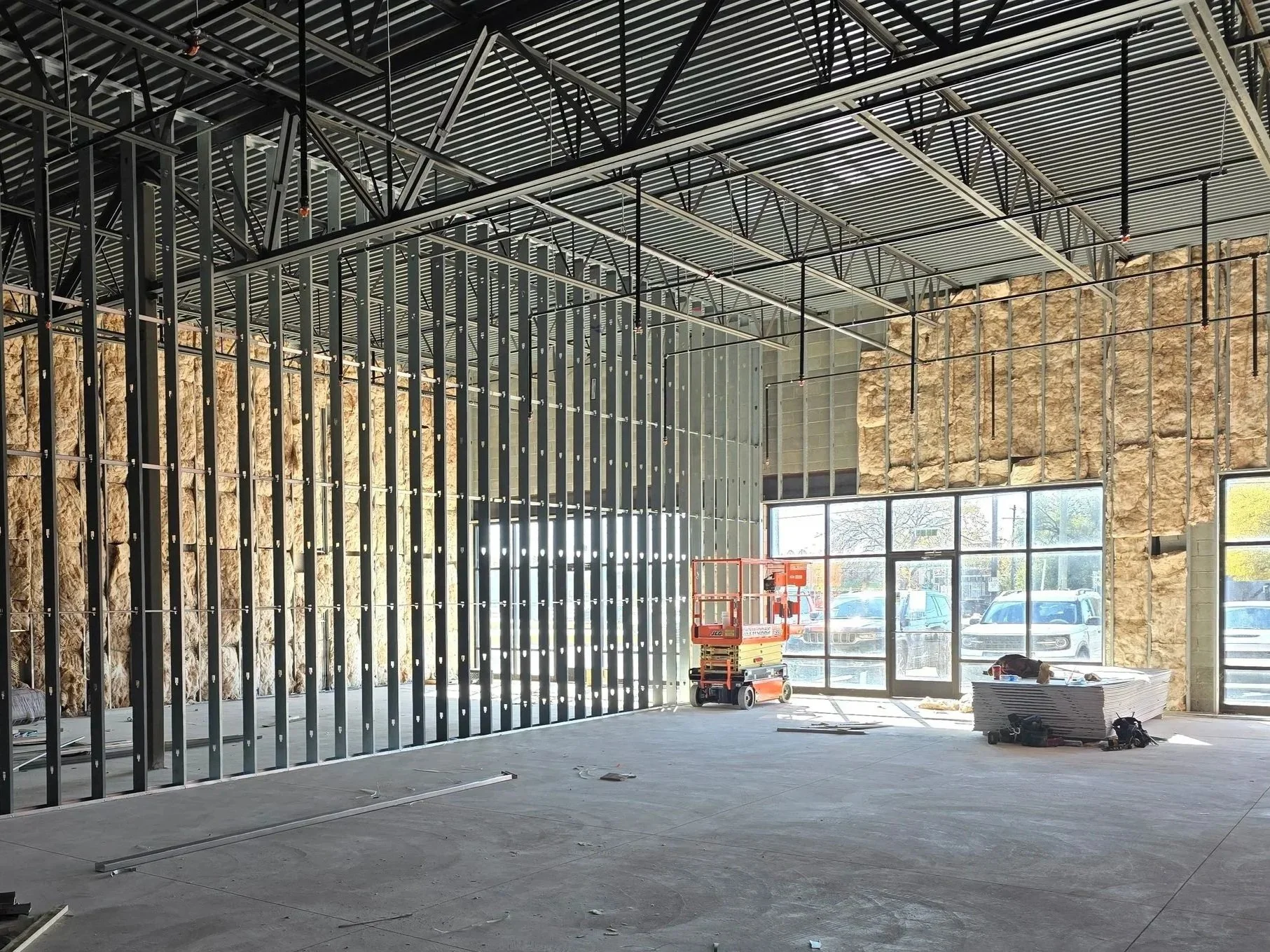 Construction site interior with exposed steel framing, insulation, large windows, and construction equipment.