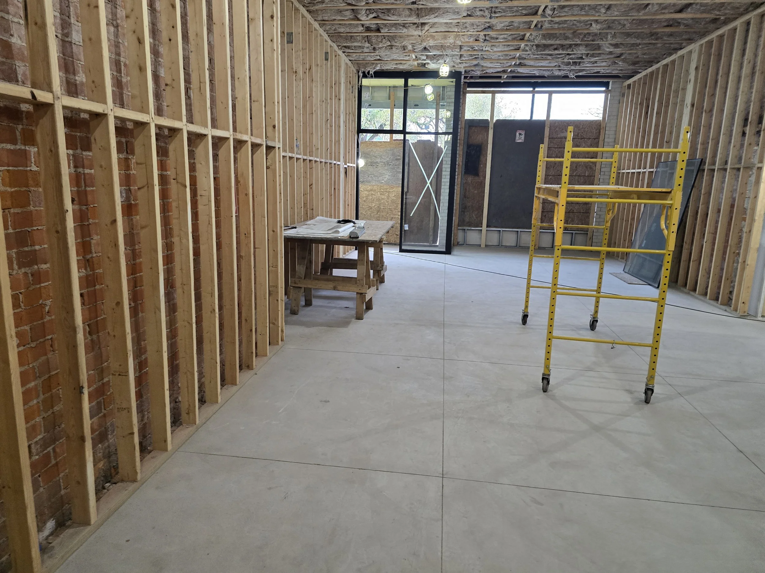 Interior of a building under construction with exposed brick wall and wooden framing, a work table with construction plans, and a yellow scaffold near large windows.