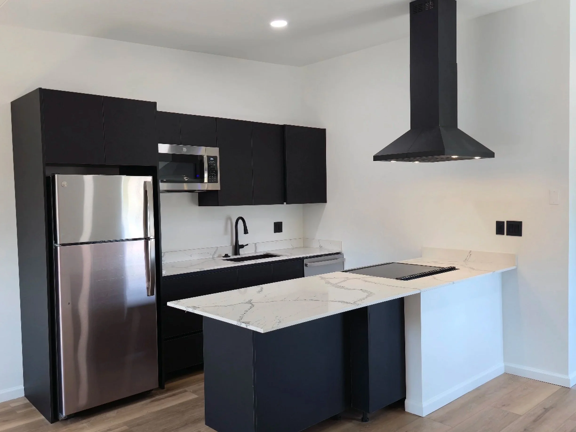 Modern kitchen with black cabinets, stainless steel refrigerator and microwave, white marble countertop, black backsplash, and a black range hood.