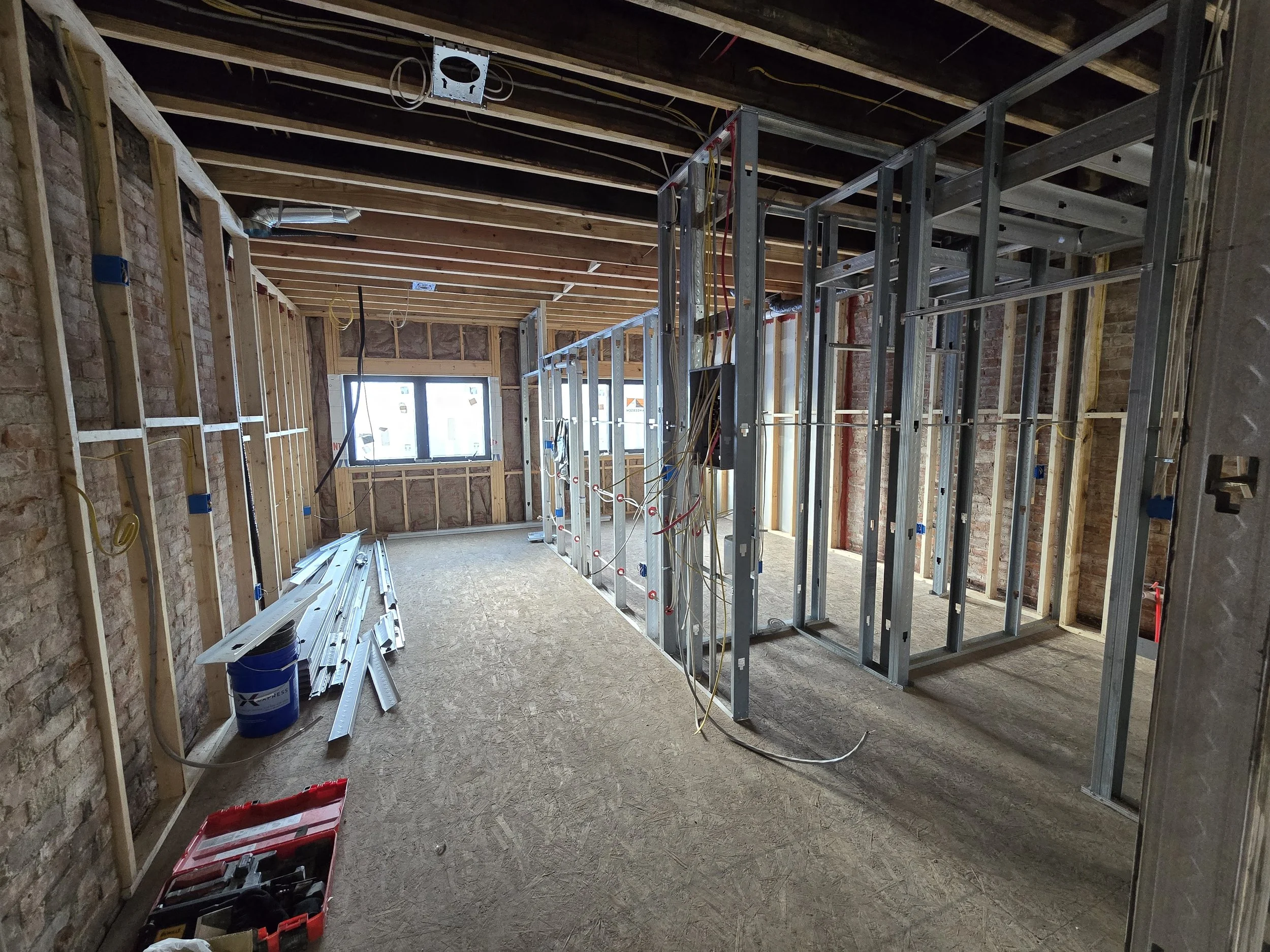 Construction site showing a room framed with metal studs, with exposed brick walls and a plywood floor. Electrical wiring and conduit are visible, with some tools and materials on the floor and leaning against the walls.