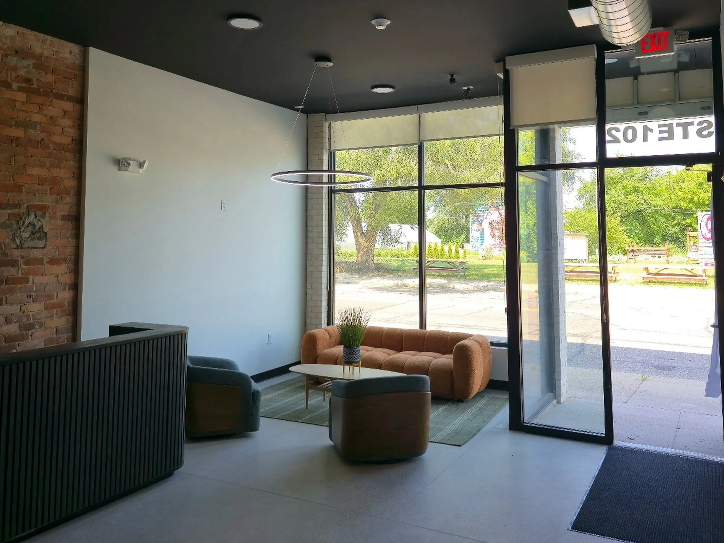Modern interior space with a large window, exposed brick wall, black ceiling, pendant lighting, and comfortable seating area with beige sofa and dark chairs.