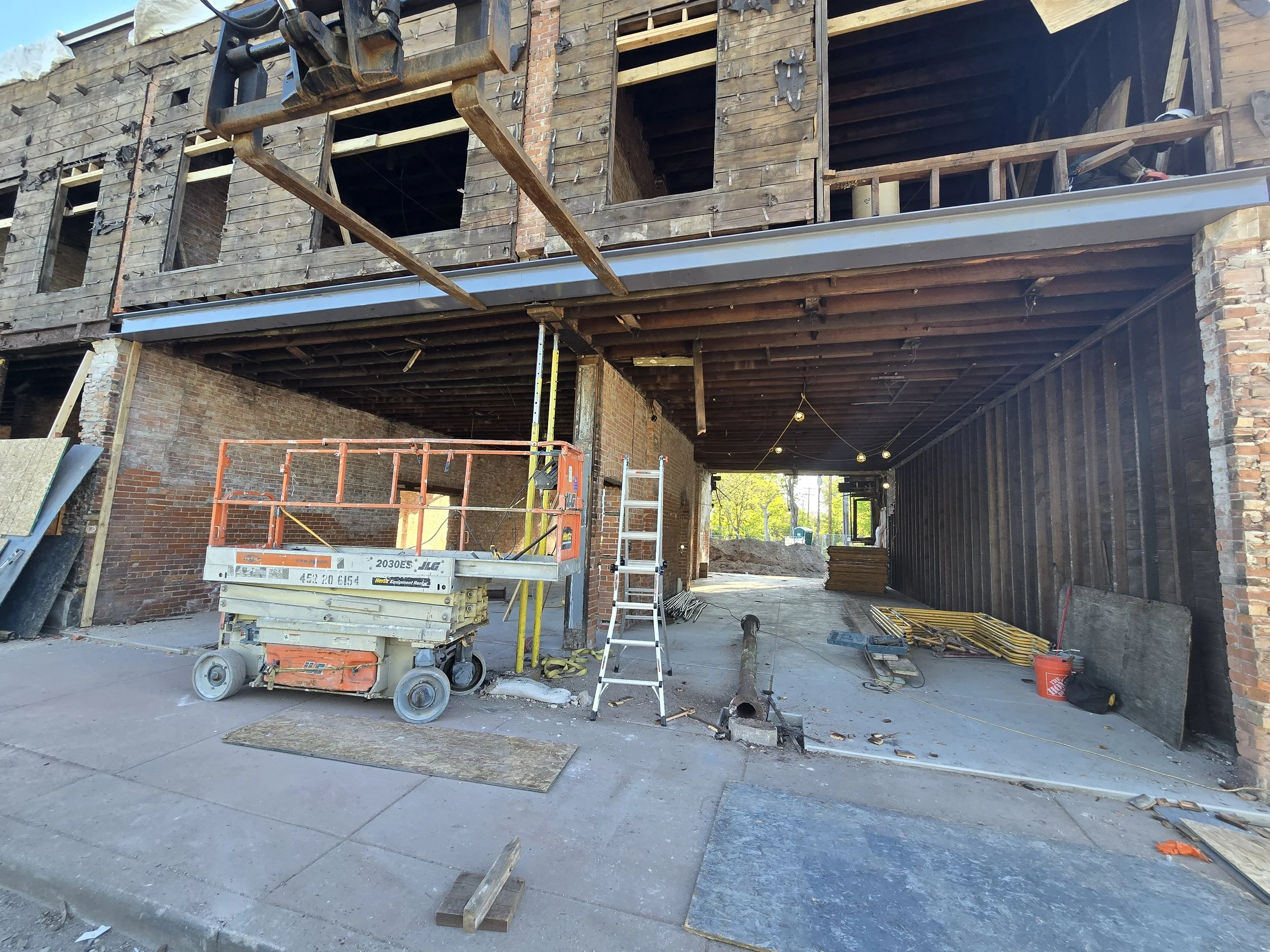 Construction site showing a building in progress with exposed brick and wooden framework, scaffolding, and construction tools and equipment like a lift, ladders, and piles of wood.