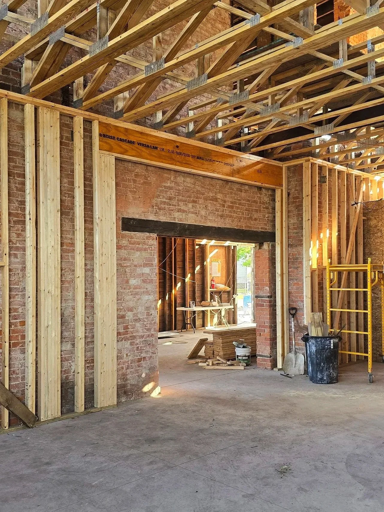 Interior of a building under construction with exposed brick walls and wooden framing for the ceiling and walls, construction tools and materials scattered around, and scaffolding in the background.