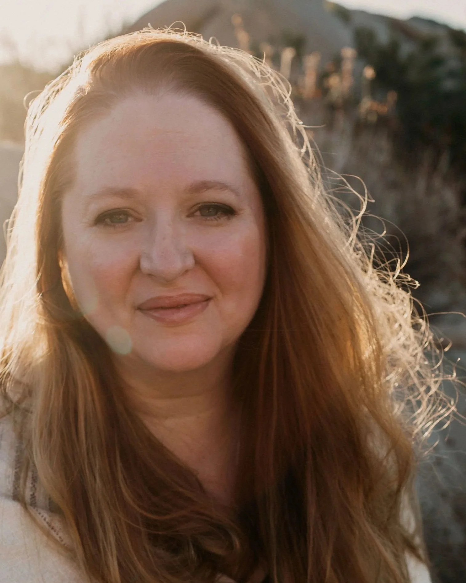 Woman smiling outdoors during golden hour, with long auburn hair and a softly blurred natural background.