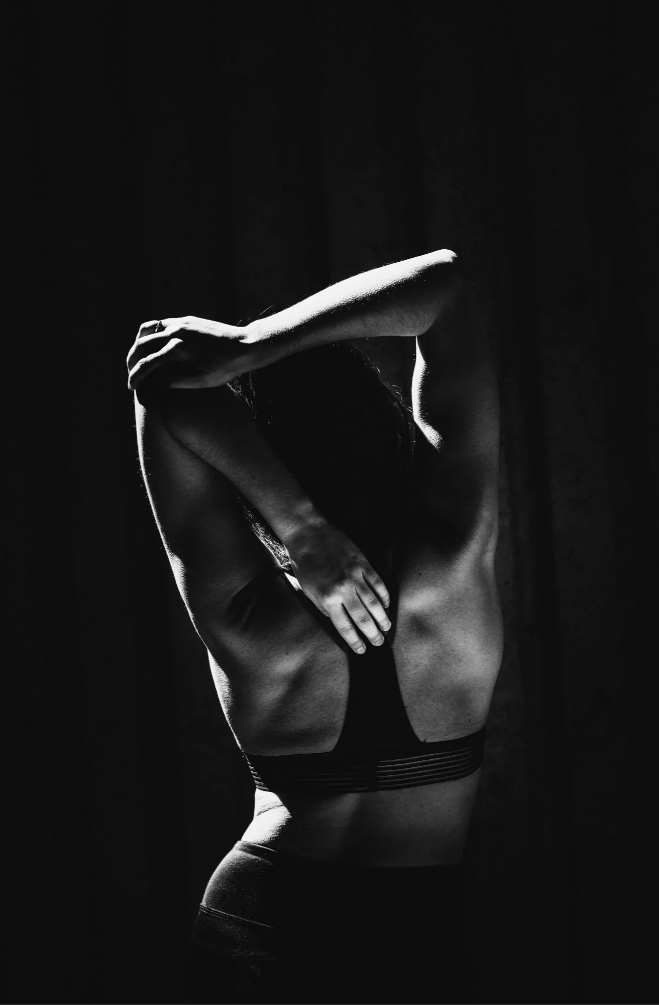 Schwarz-weiß-Fotografie eines nackten Menschen, der sich in einer Pose mit erhobenen Armen und gekreuzten Beinen befindet, vor einem dunklen Hintergrund.