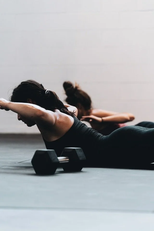 Zwei Frauen machen Bauchmuskelübungen auf einer Yogamatte, mit Hanteln in der Nähe, in einem Fitnessstudio.