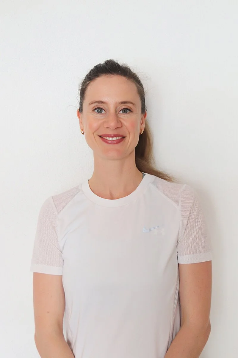 Eine lächelnde Frau mit braunem Haar, die einen weißen Sport-T-Shirt trägt, steht vor einer weißen Wand.