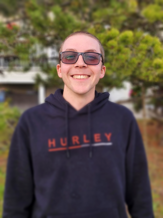 Smiling young man wearing sunglasses and a black Hurley hoodie, standing outdoors with green trees and a house in the background.
