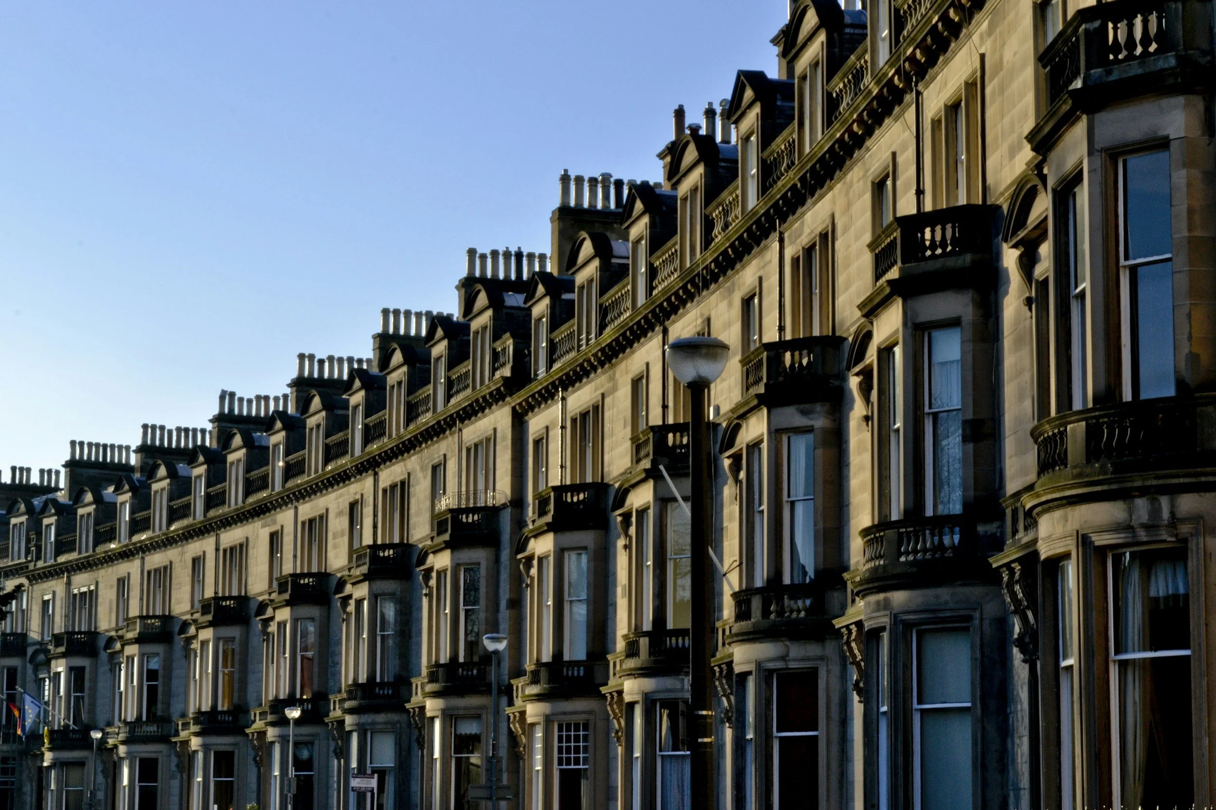 A row of historic beige stone apartment buildings with balconies, large windows, and chimney stacks, set against a clear blue sky.