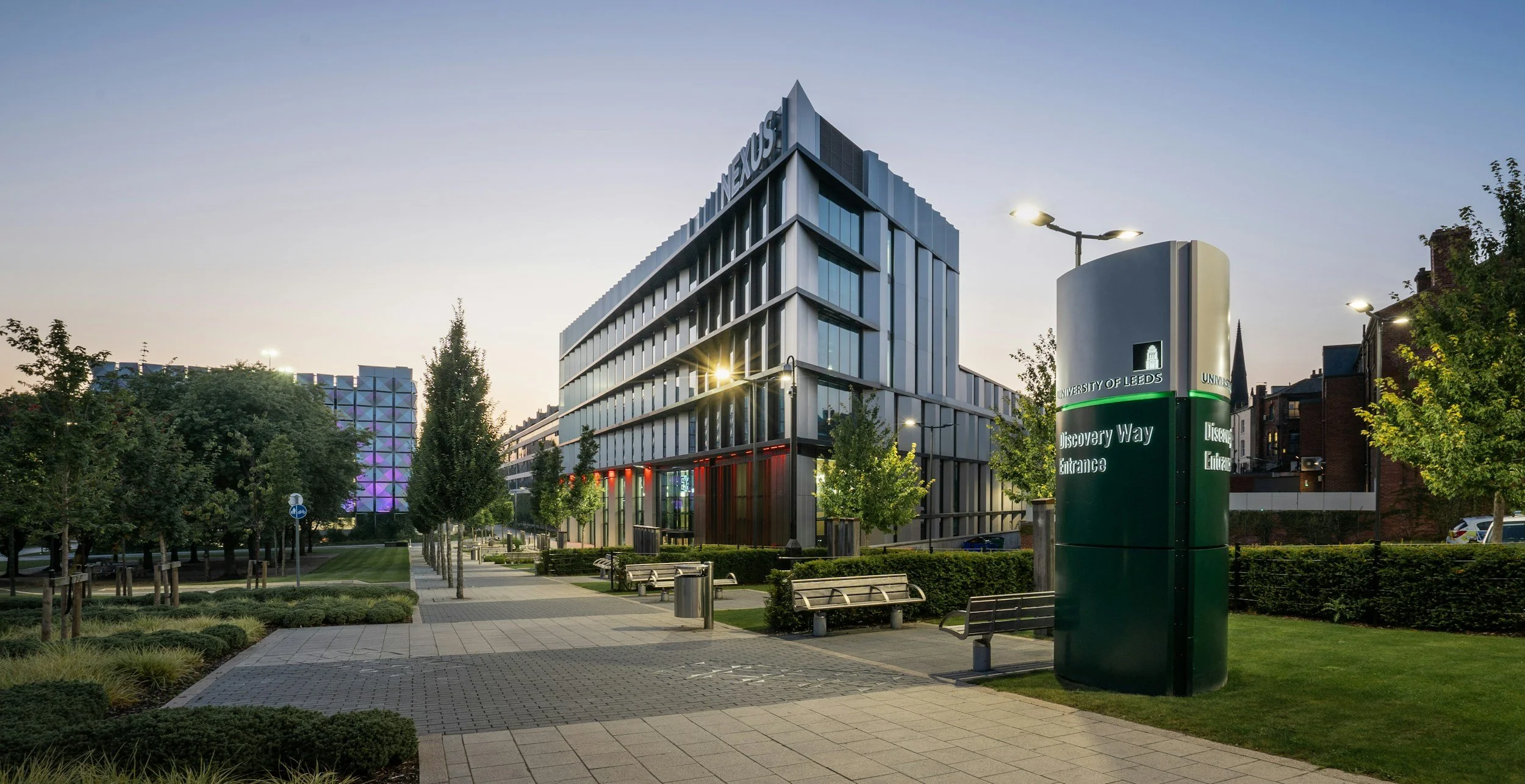 Modern university building with glass windows, trees, benches, and a sign that reads 'University of Leeds Discovery Way Entrance'.