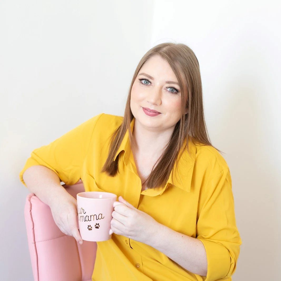 A woman with light skin and straight brown hair, wearing a yellow shirt, sitting on a pink chair, holding a pink coffee mug that says 'Our Mama' with paw prints, against a plain white background.