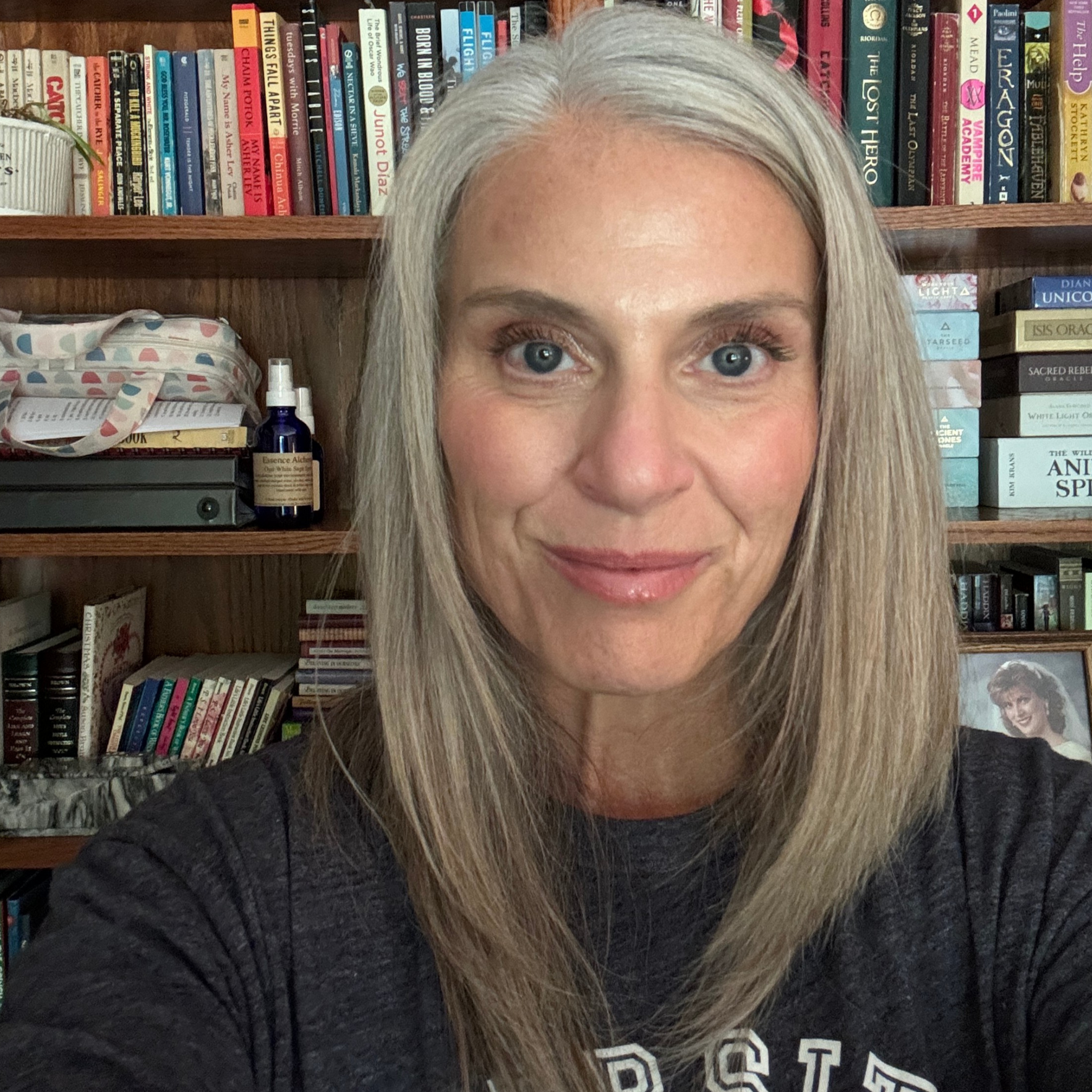 A woman with long blonde hair, blue eyes, and light skin taking a selfie in front of a bookshelf filled with various books. She is wearing a black shirt with white text on it and smiling slightly.
