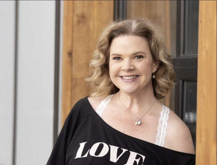 A woman with blonde, curly hair smiling at the camera, wearing a black top with the word "LOVE" on it, layered with a white lace strap under her shoulder, standing in front of a wooden and black-paneled wall.