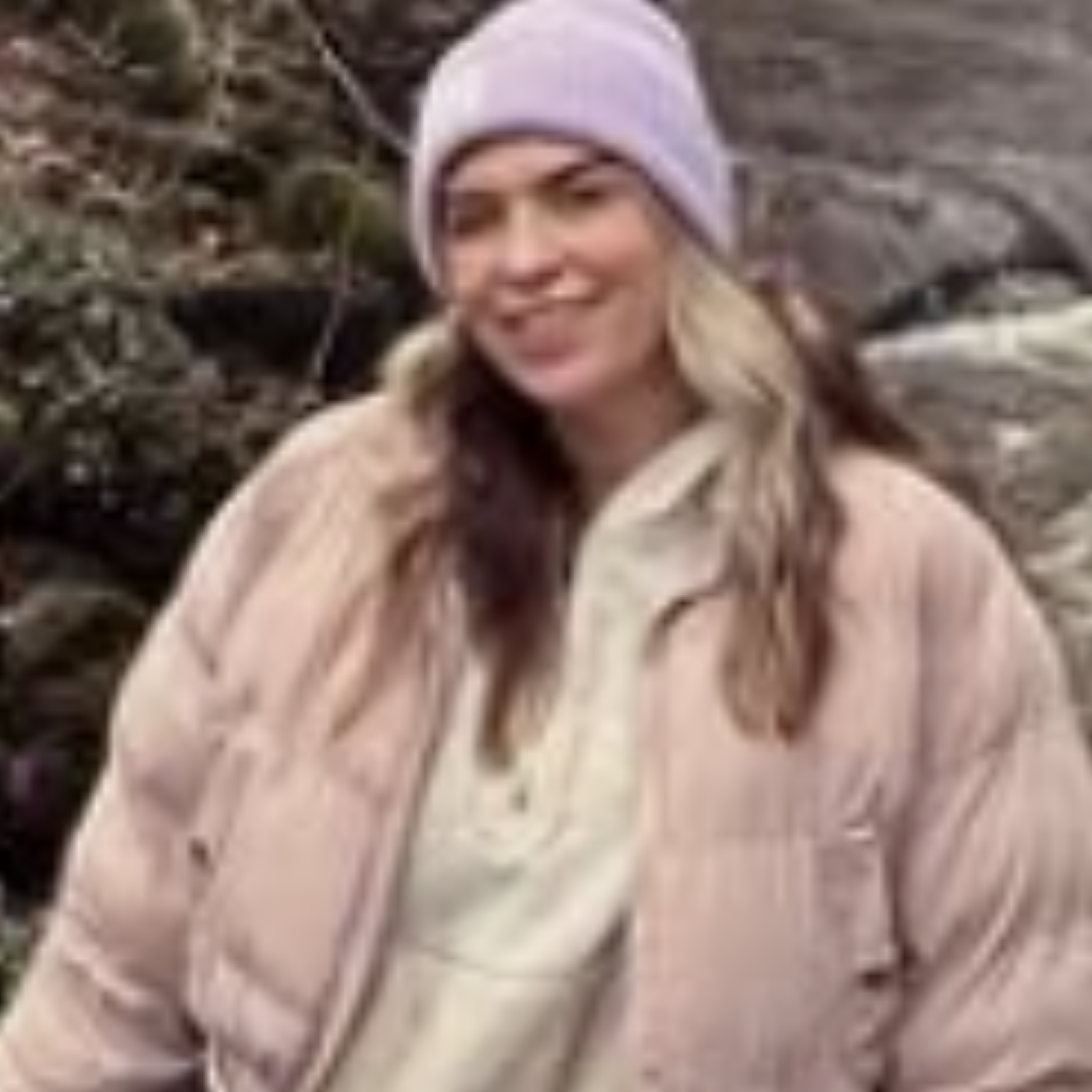 A woman smiling outdoors, wearing a white beanie, with a background of rocks and vegetation.