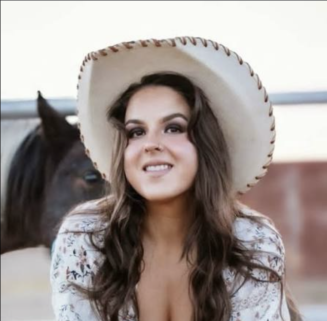 Woman with long wavy hair wearing a large cowboy hat and floral shirt, standing next to a horse.