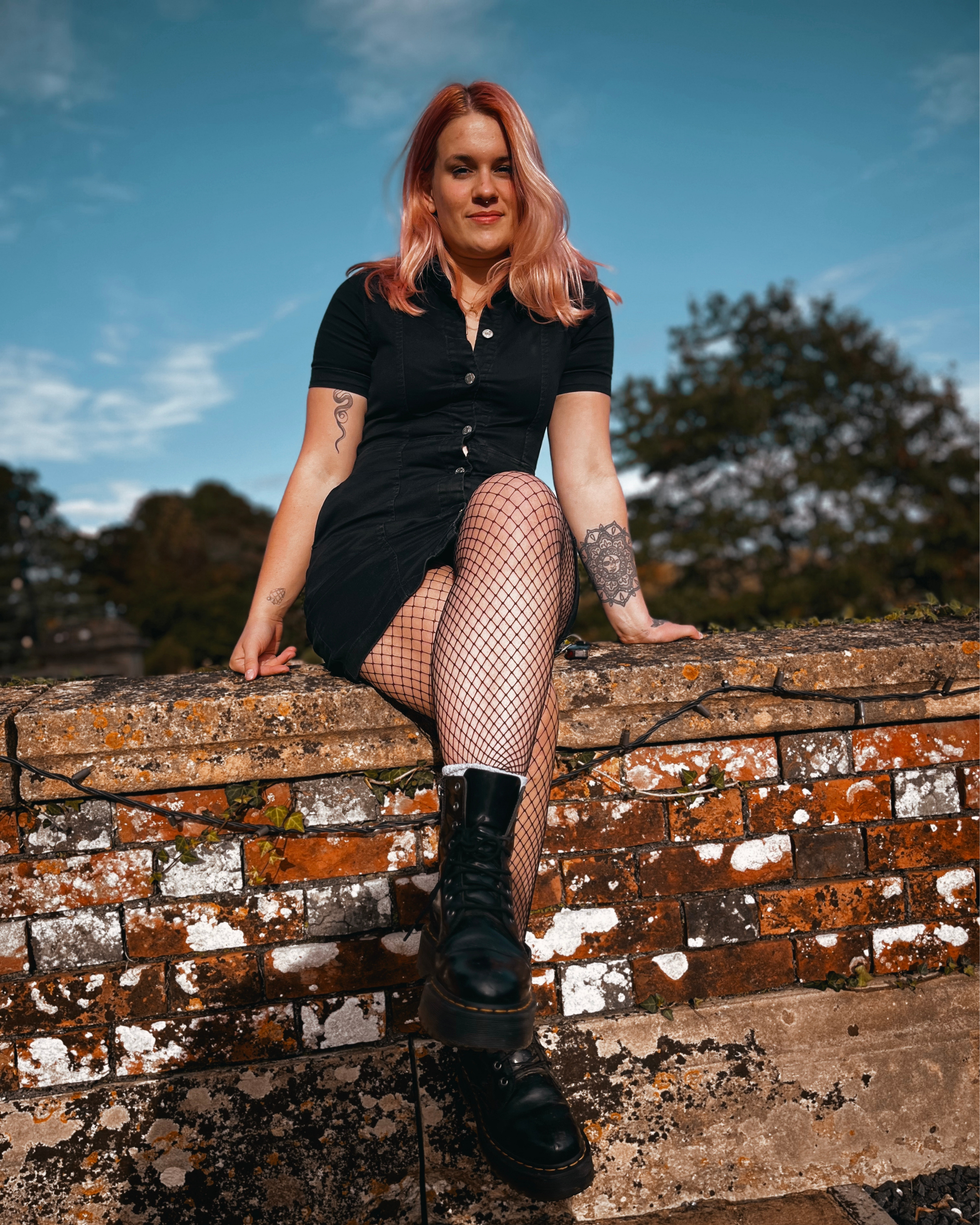 A woman with pink hair and tattoos, wearing a black dress, fishnet stockings, and black combat boots, sitting on a brick wall outdoors during the evening with a partly cloudy sky and trees in the background.