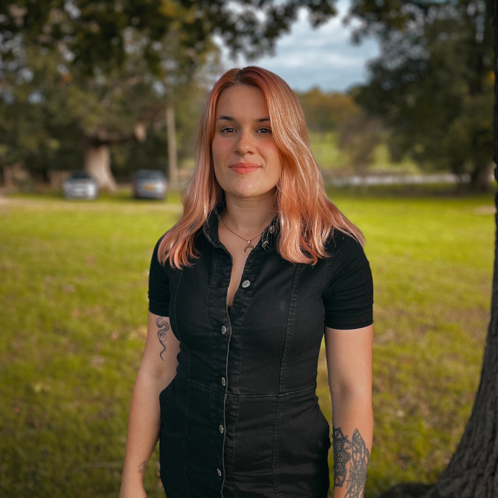 A young woman with shoulder-length pinkish-blonde hair, wearing a black button-up shirt with short sleeves, standing outdoors on a grassy area with trees and parked cars in the background. She has tattoos on her right arm and is smiling gently.