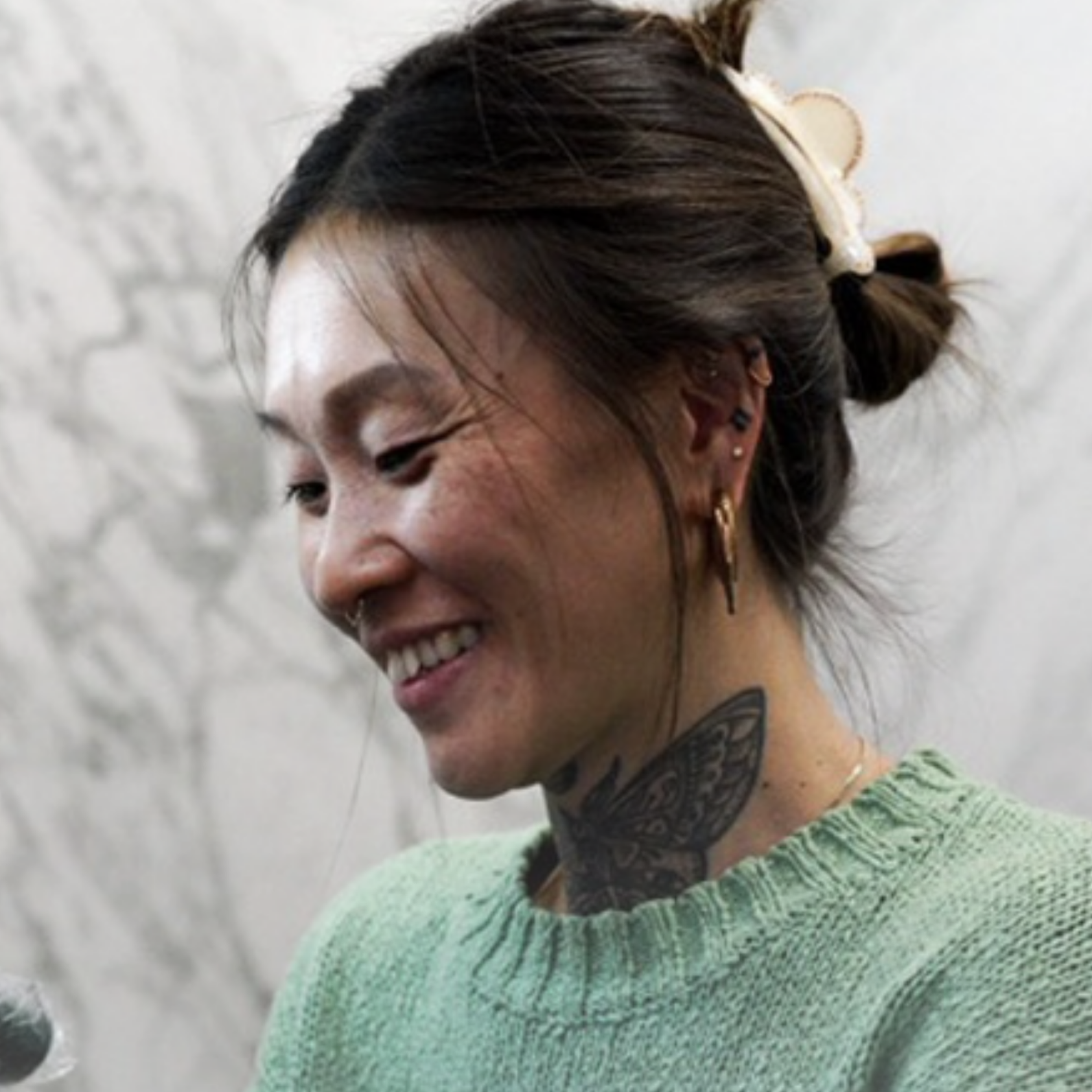 A woman with dark brown hair styled in a bun secured with a large cream-colored hair clip, showing multiple earrings, smiling with eyes closed, wearing a green knitted sweater, standing against a textured white wall.