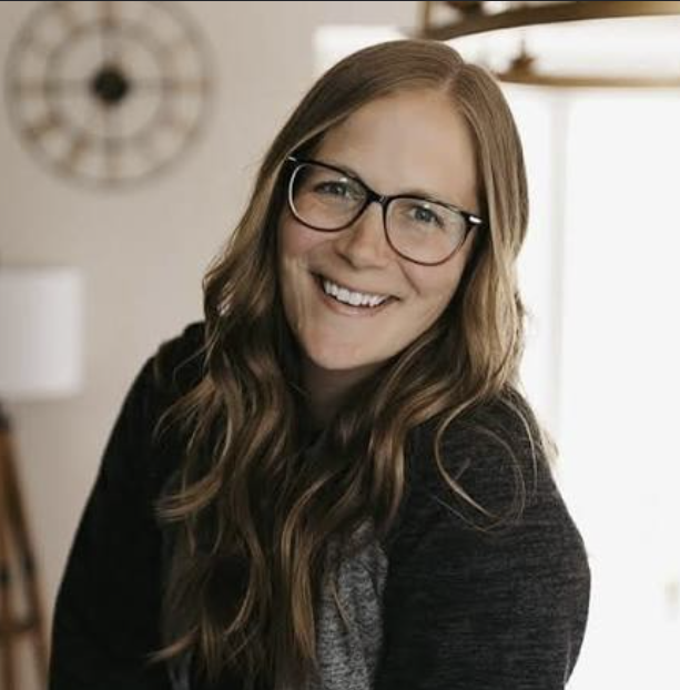 A smiling woman with long, wavy brown hair and glasses, sitting indoors near a window.