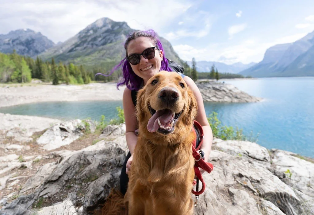 A woman with purple hair and sunglasses smiling with her dog by a lake in mountainous terrain.