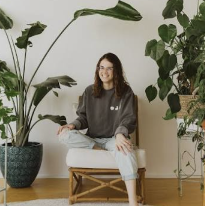 A woman with glasses and curly hair sitting on a wooden chair, surrounded by large indoor plants in a well-lit room.