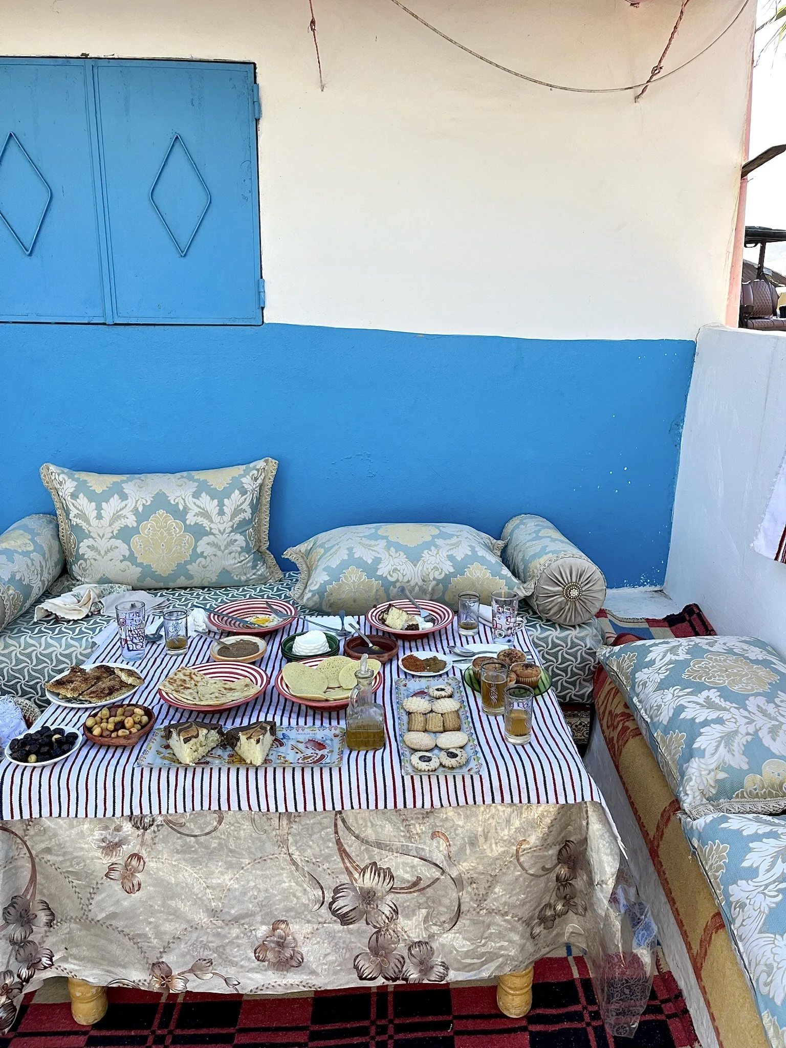 A table set for a meal, displaying a variety of foods including bread, pastries, snacks, and drinks, with pillows and decorated walls in the background.