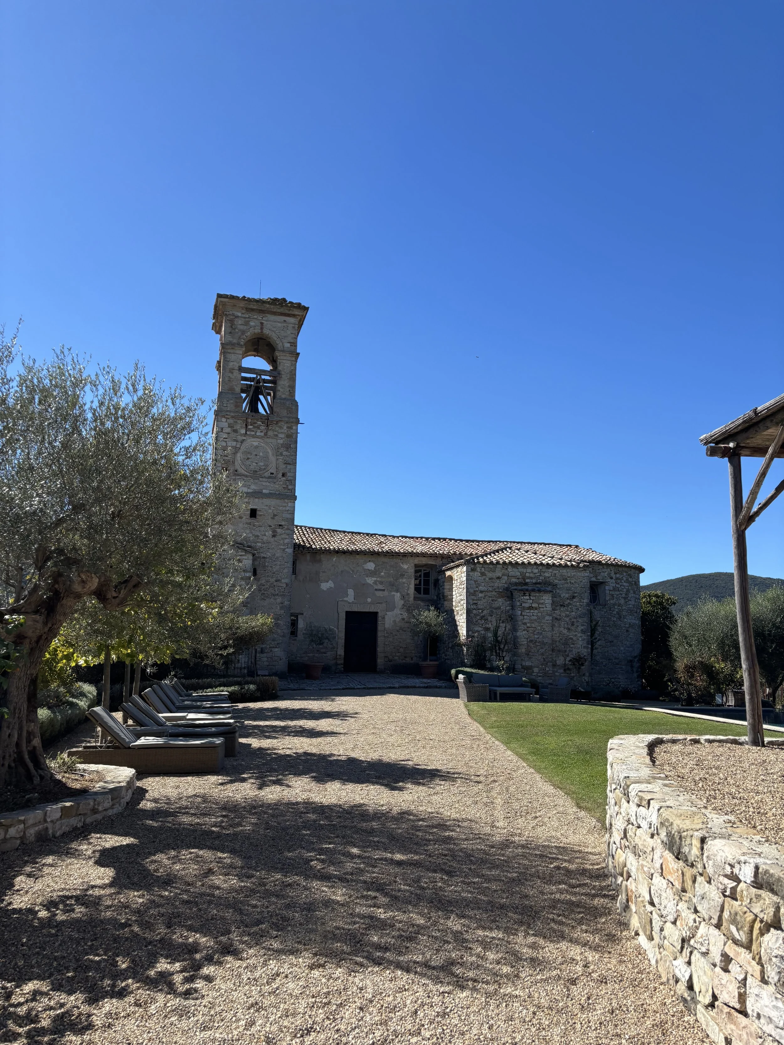 A historic stone church with a bell tower, surrounded by a courtyard with lounge chairs, trees, and a stone wall under a clear blue sky.