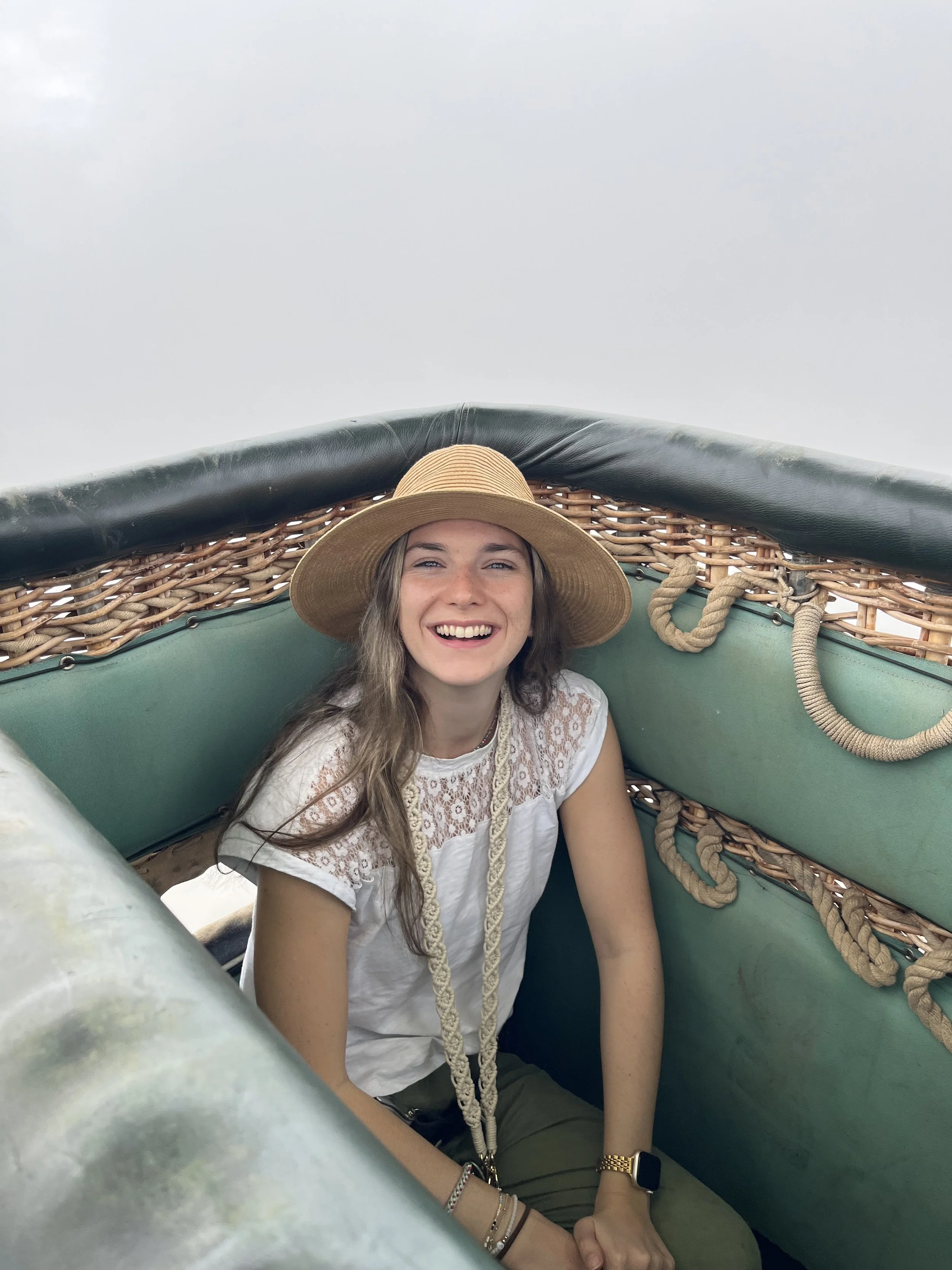 Young woman with light skin and long hair smiling, wearing a wide-brimmed straw hat and a white embroidered top, sitting inside a bamboo and canvas hot air balloon basket outdoors during the day.