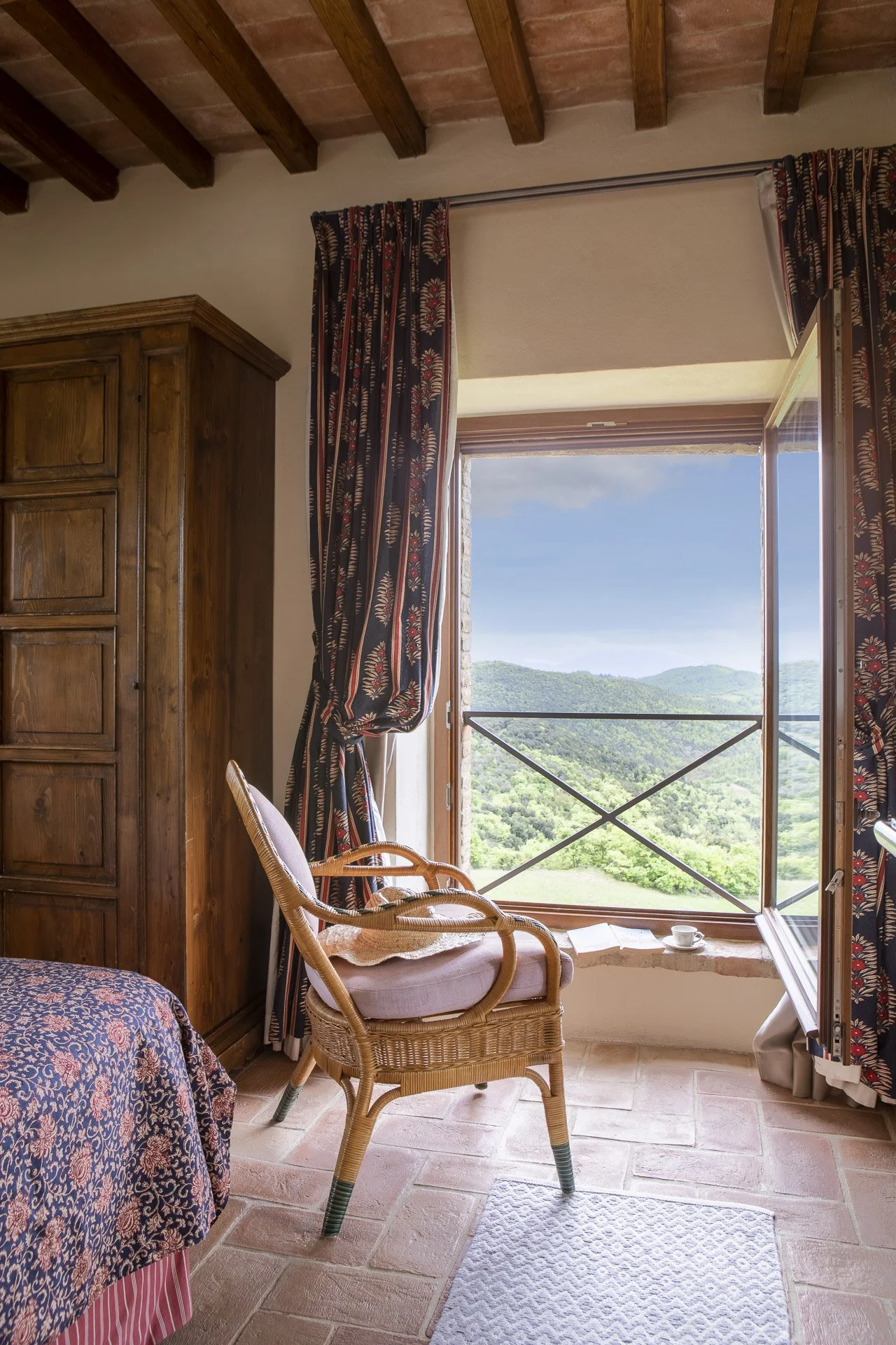 Room with a large open window showing green hills and blue sky, wooden ceiling beams, a wooden wardrobe, a wicker chair with a cushion, a bed with a floral bedspread, and patterned curtains.