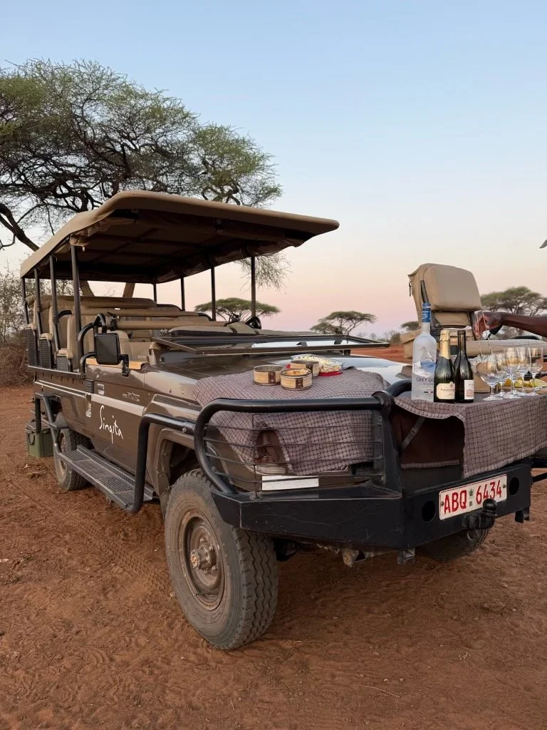 A safari vehicle set up for a meal with a table covered with a tablecloth, wine bottles, glasses, and snacks, parked on a dirt path with trees and a clear sky in the background.