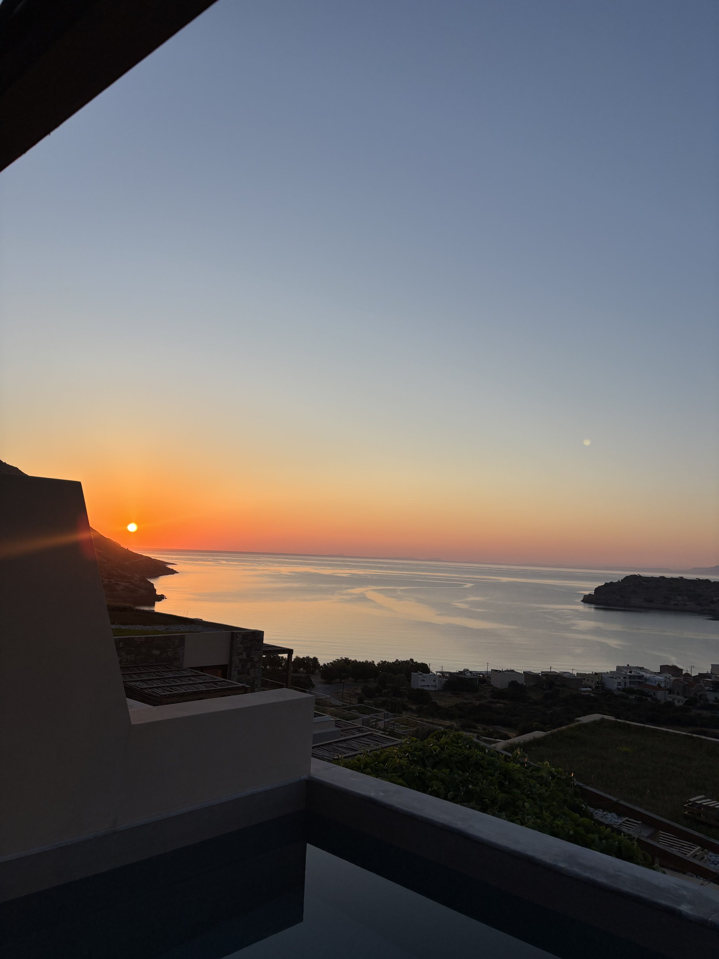Sunset over the ocean seen from a balcony with modern white railings and buildings, clear sky with a small bright moon.