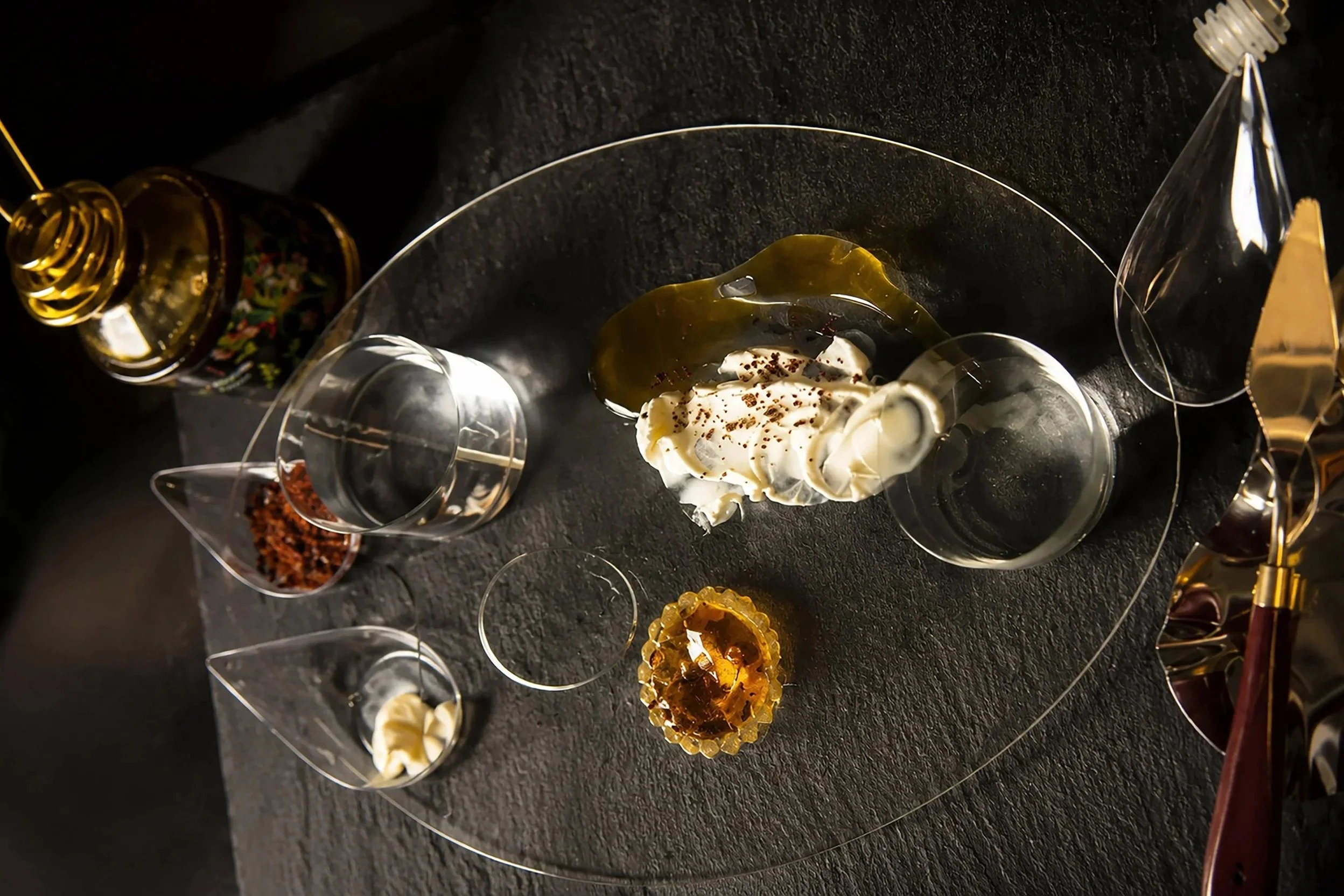 Several small clear plastic cups with various candies and treats arranged on a black surface, some with desserts, a bottle of alcohol, and decorative gold and black items.