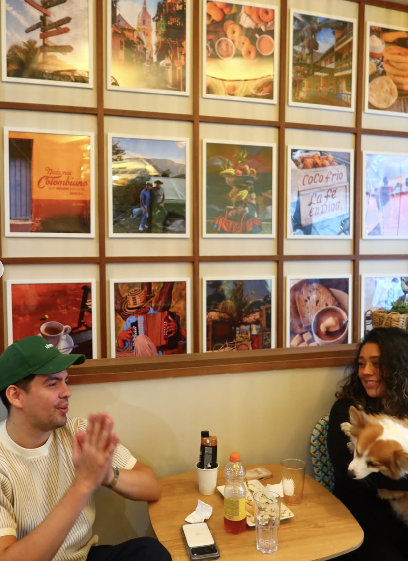 Dos personas en una mesa de un café, una mujer con un perro y un hombre con gorra verde, con fotografías enmarcadas en la pared atrás que muestran escenas de Colombia y comida típica.