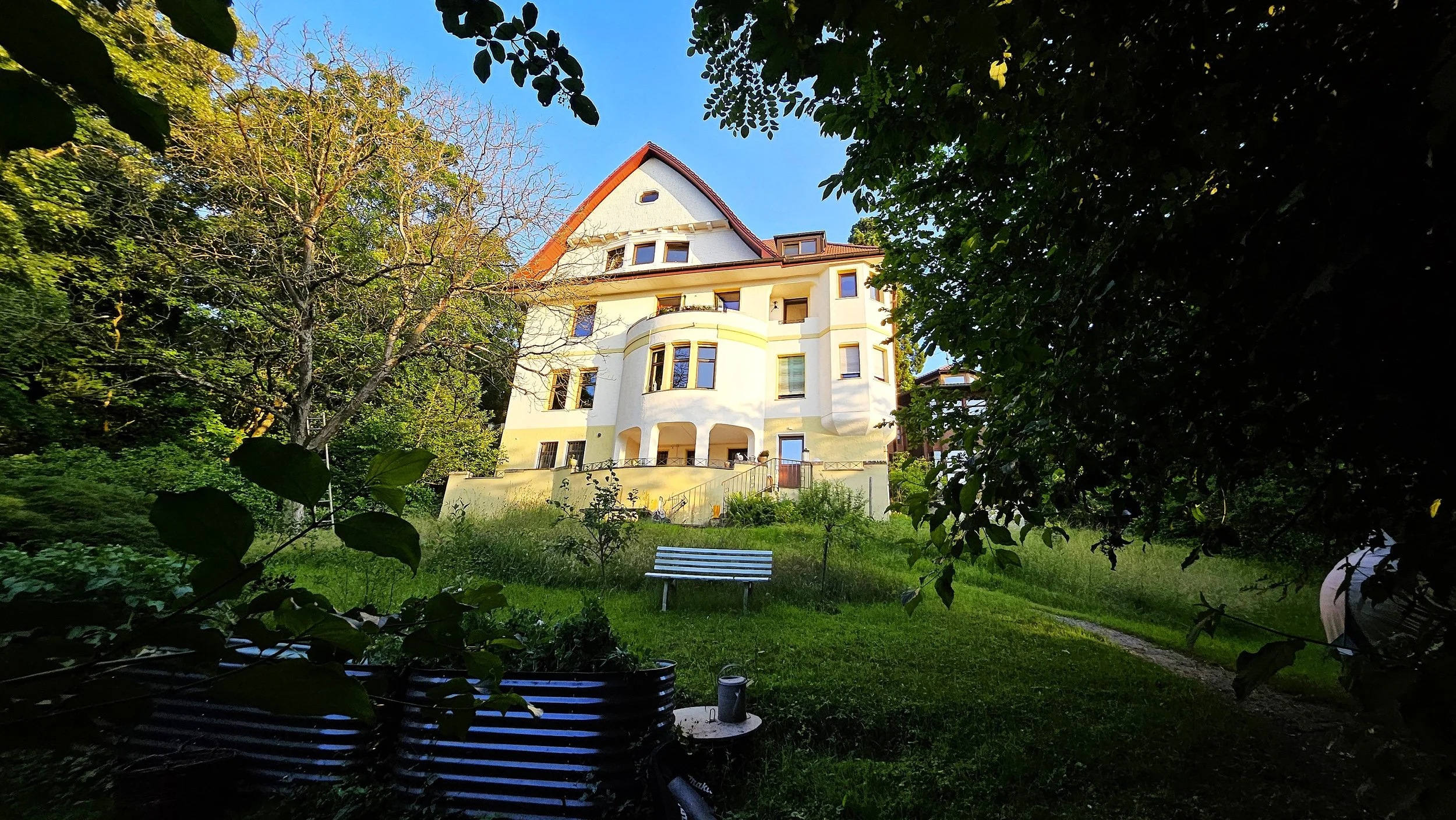 Ein mehrstöckiges Haus mit weißen Wänden und rotem Dach, umgeben von Bäumen und grünem Garten, im Sonnenlicht.