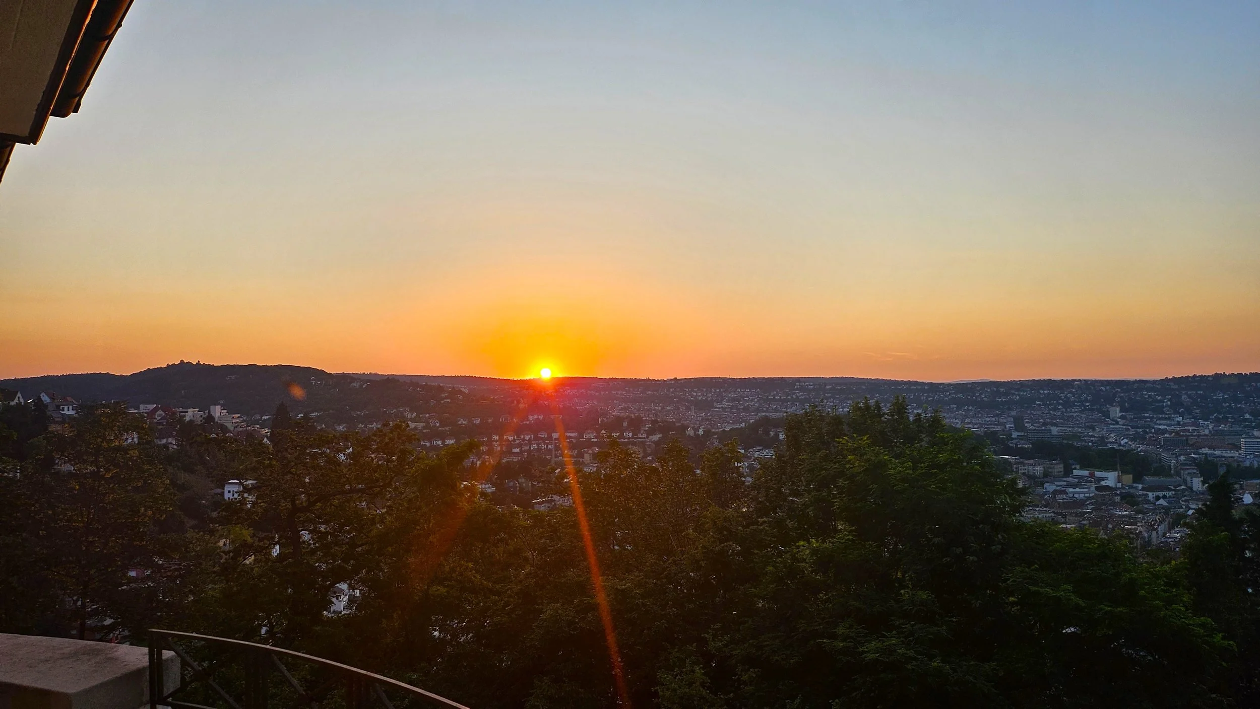 Blick auf eine Stadt bei Sonnenuntergang über Bäume hinweg, mit orangefarbenem Himmel und der Sonne am Horizont.
